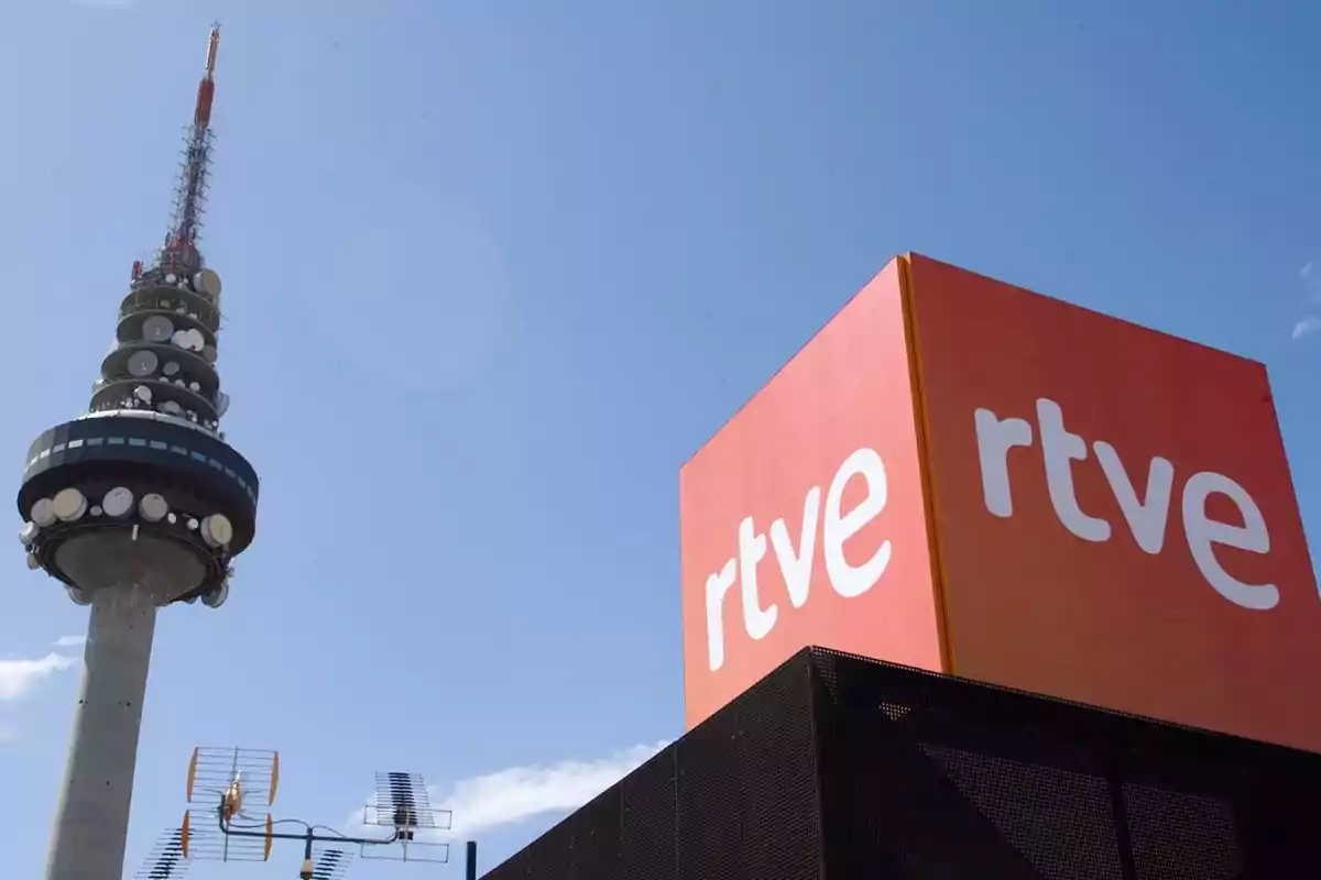 Torre de telecomunicaciones junto a un edificio con el logotipo de RTVE bajo un cielo despejado.