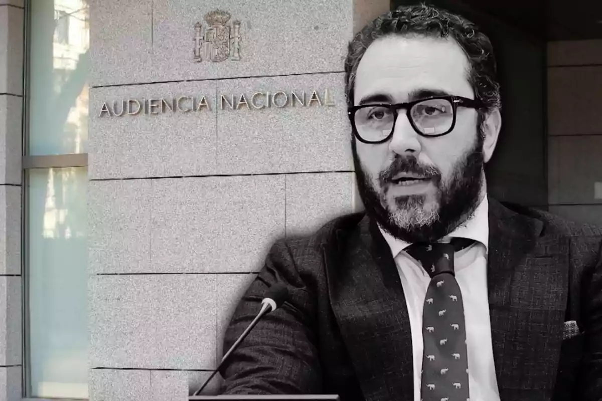 Hombre con barba y gafas hablando frente a un micrófono con el edificio de la Audiencia Nacional de fondo.