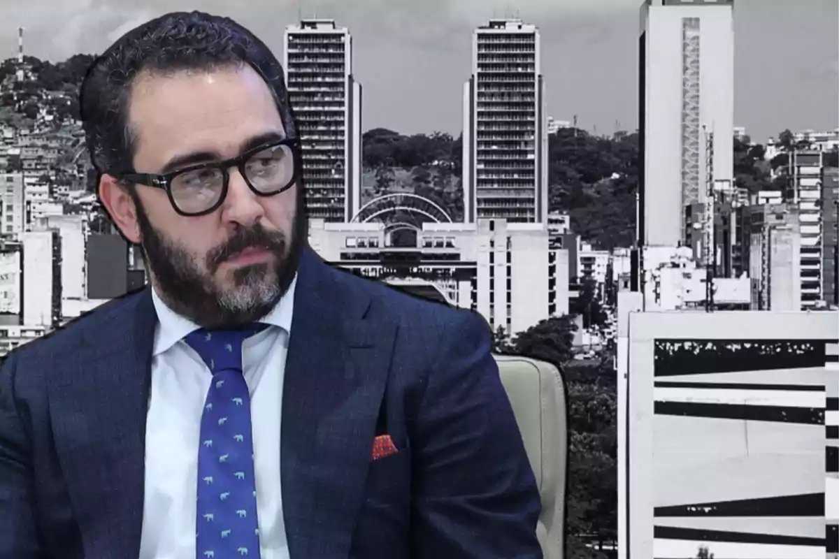 Un hombre con barba y gafas, vestido con traje y corbata azul, sentado frente a un fondo de edificios en blanco y negro.