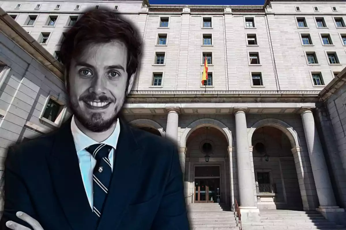 Un hombre sonriente con traje y corbata frente a un edificio gubernamental con una bandera española.