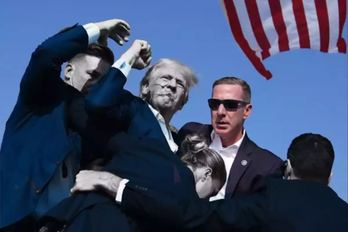 A group of people in dark suits surround an older man with his fist raised, while an American flag flutters in the background.