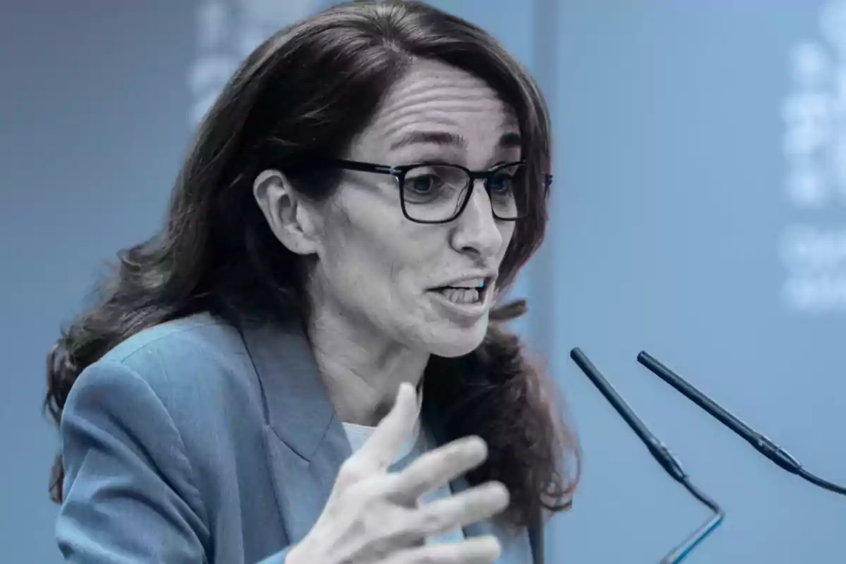 Una persona con gafas y cabello largo hablando en un micrófono durante una conferencia.