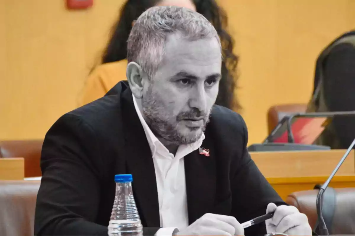 A man with a beard and gray hair is sitting at a table, holding a pen, with a bottle of water in front of him.