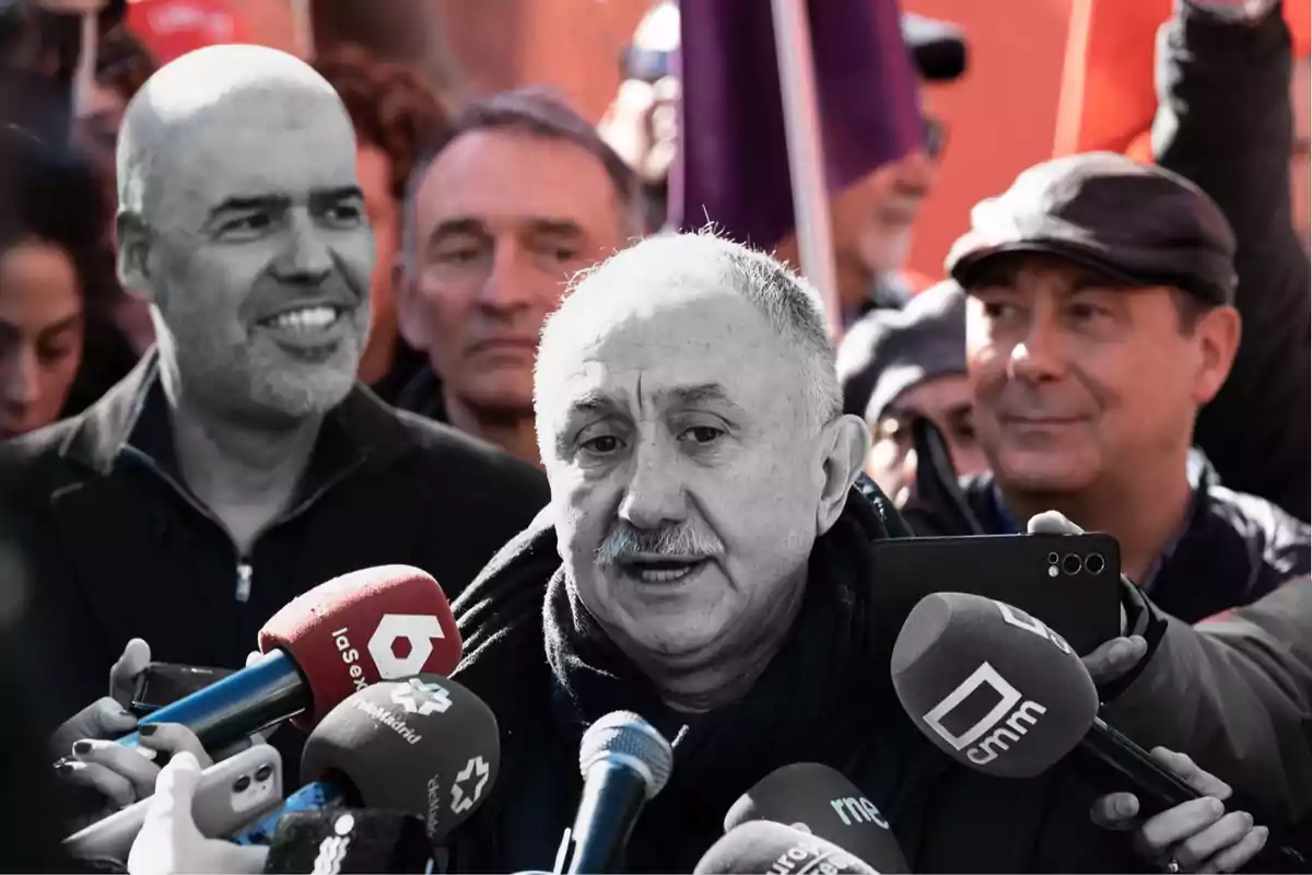 An elderly man speaks in front of several microphones from different media outlets while surrounded by people at an outdoor event.