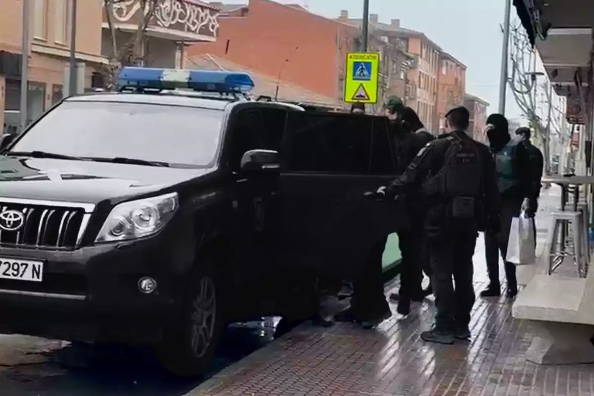 Un grupo de personas vestidas de negro junto a un vehículo con luces azules estacionado en una acera urbana.