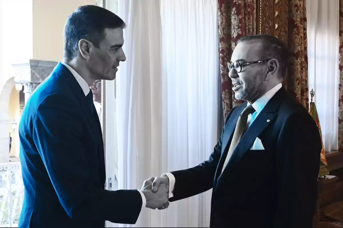 Two men in suits shake hands in a room with white curtains and elegant decor.