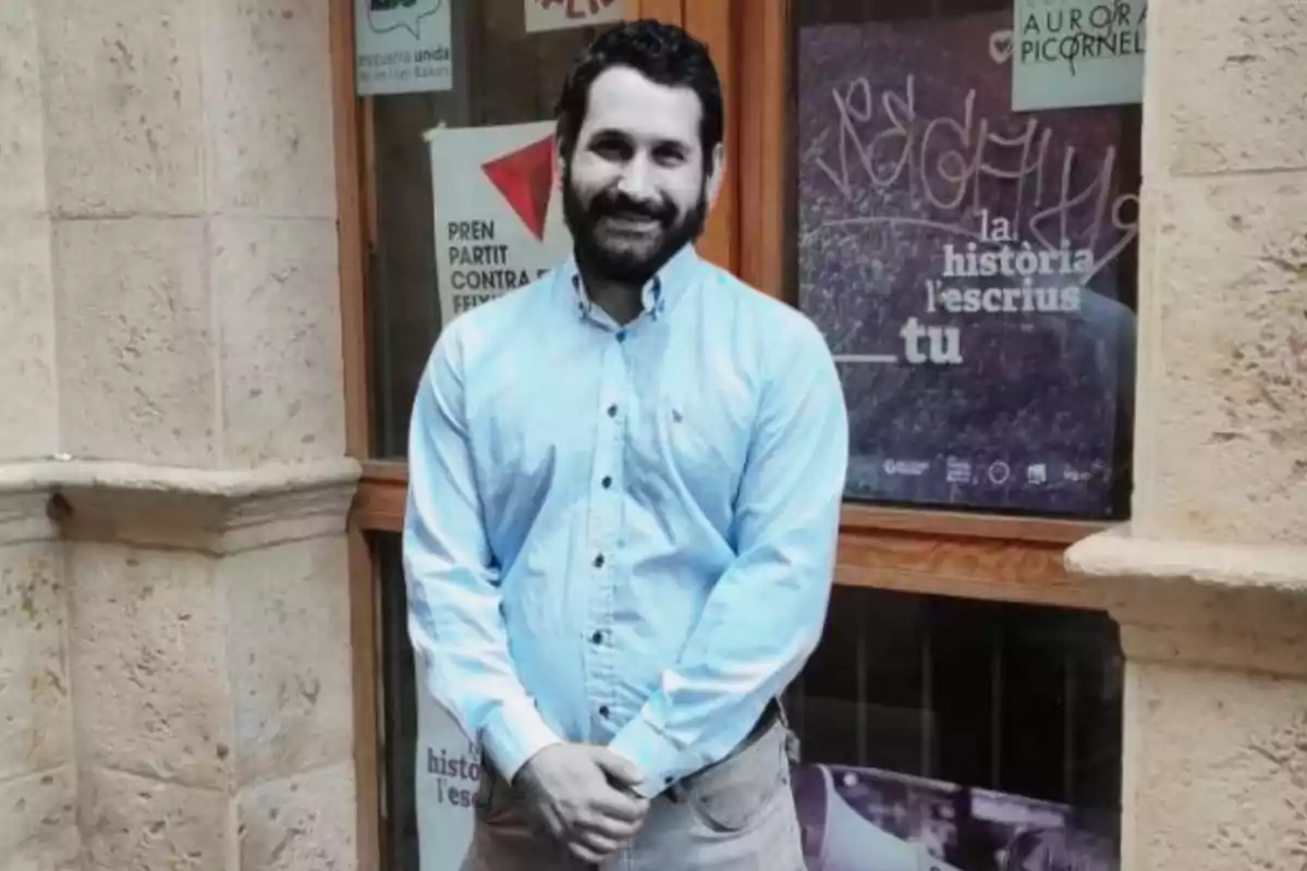 A man in a light blue jersey posing in front of a stone wall and a window with posters.