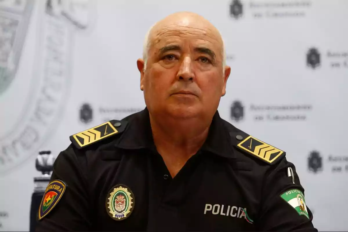 Un oficial de policía con uniforme negro y emblemas en los hombros y pecho está sentado frente a un fondo con logotipos.