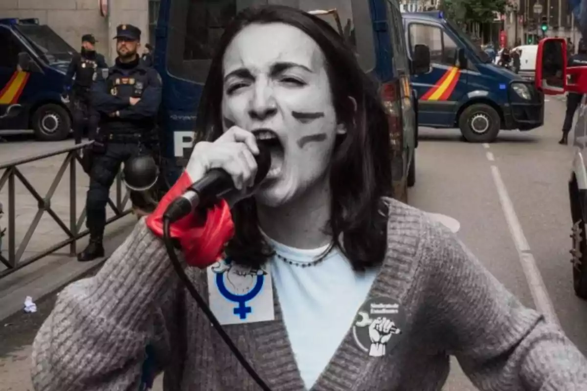 A person with face paint holds a microphone while protesting in a street, with police vehicles and officers in the background.