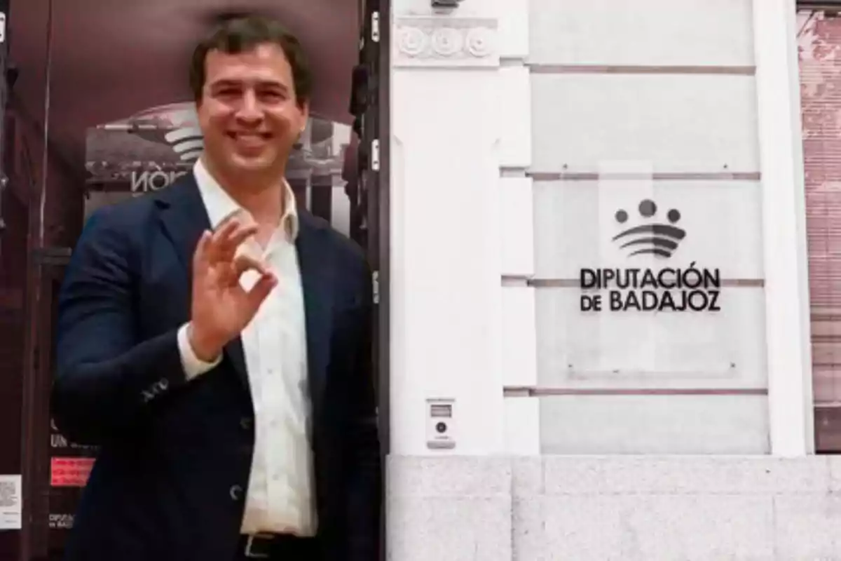 A smiling man in a dark suit gestures with his hand in front of a building with a sign that says "Legislature de Badajoz."