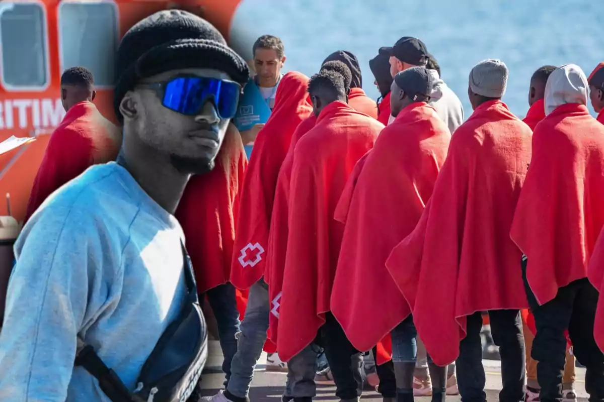 Un grupo de personas cubiertas con mantas rojas se encuentra de pie cerca de una embarcación, mientras un hombre con gafas de sol y gorro negro aparece en primer plano.