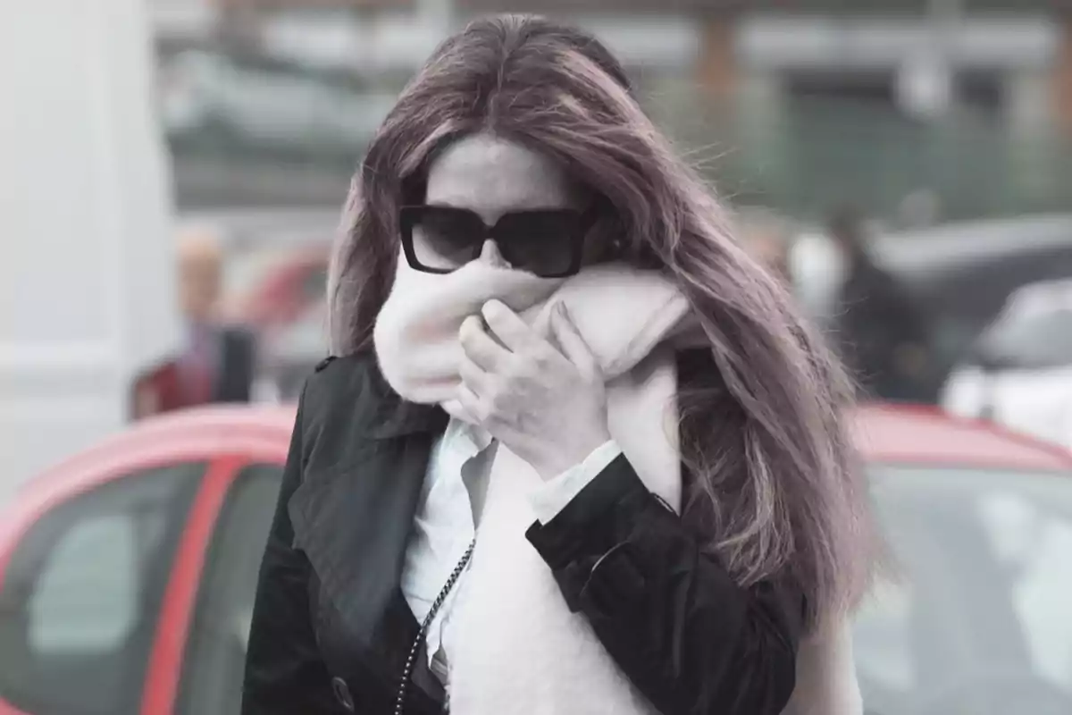 Una persona con gafas de sol y bufanda cubriendo parte de su rostro, caminando al aire libre.