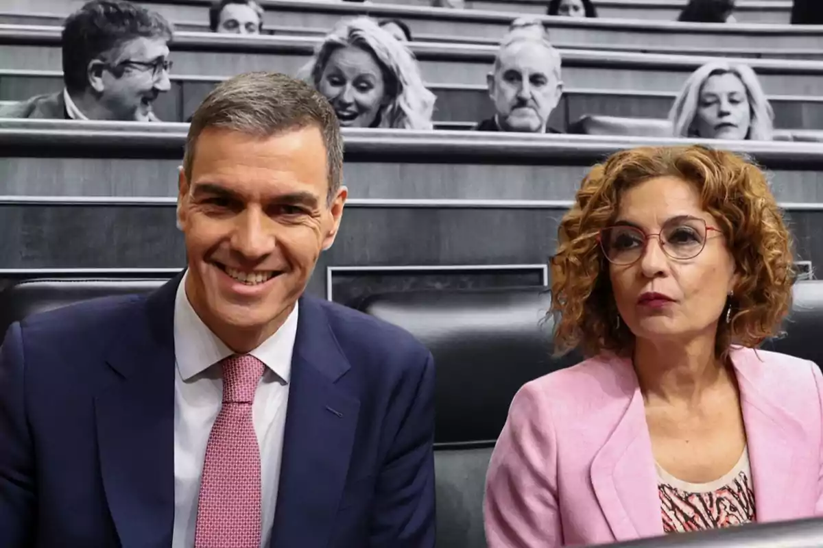 Dos personas sentadas en un entorno formal con otras personas en el fondo.