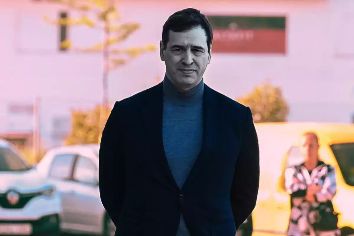 Hombre de cabello corto y oscuro vistiendo un suéter gris y saco azul, de pie al aire libre con autos y una persona desenfocada al fondo