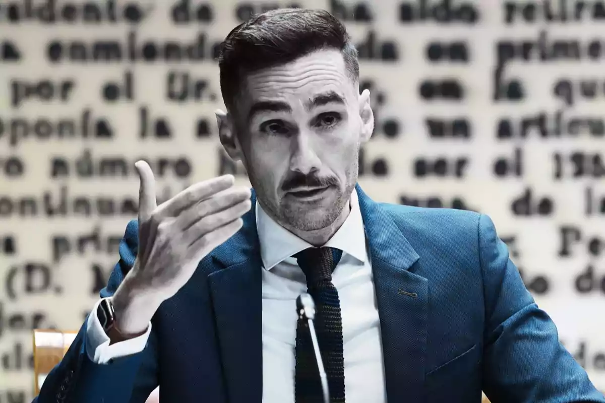 Hombre de cabello corto y barba recortada usando traje azul y corbata, gesticulando con la mano mientras habla frente a un fondo desenfocado con texto en blanco y negro