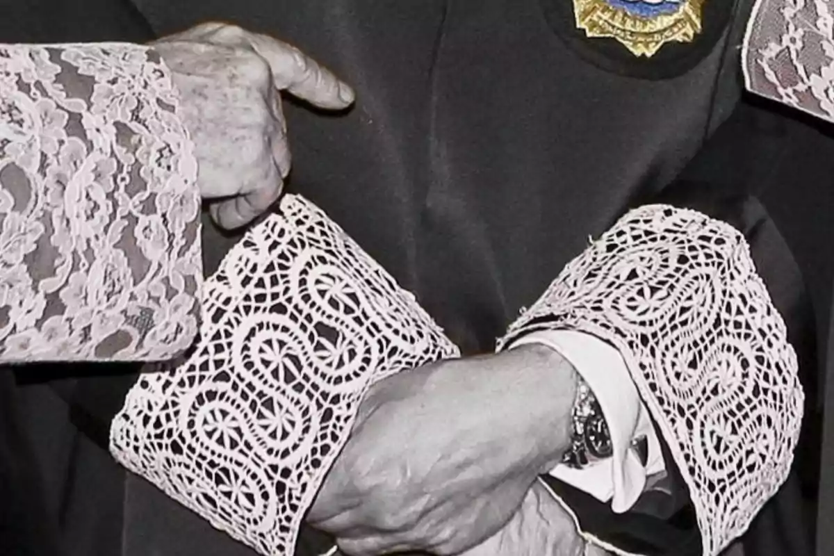 Manos de una persona mayor con puños de encaje blanco sobre una toga negra con un escudo bordado Manos de una persona mayor con puños de encaje blanco sobre una toga negra con un escudo bordado