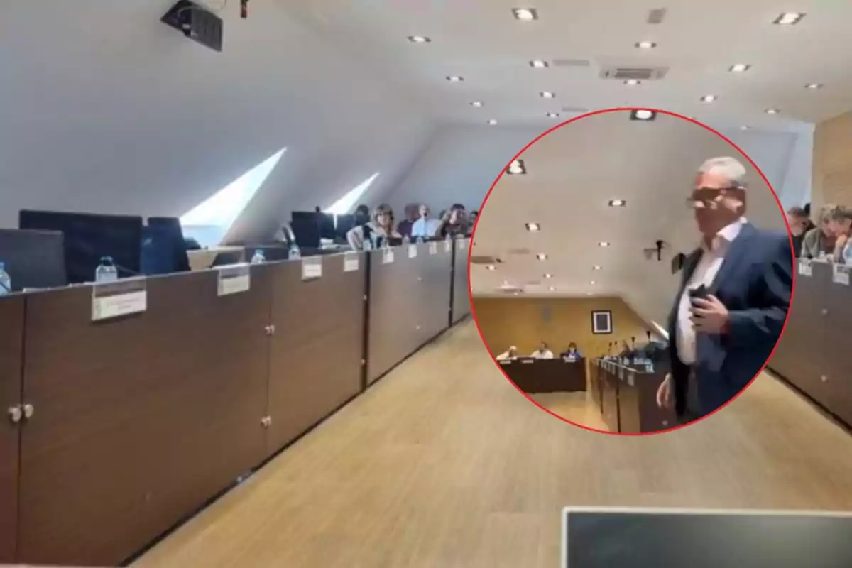 Un hombre de traje camina en una sala de reuniones con varias personas sentadas en escritorios y botellas de agua sobre la mesa