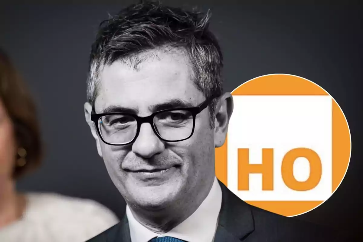 Hombre de cabello corto y canoso con gafas y traje oscuro frente a un fondo oscuro y un círculo naranja con las letras HO