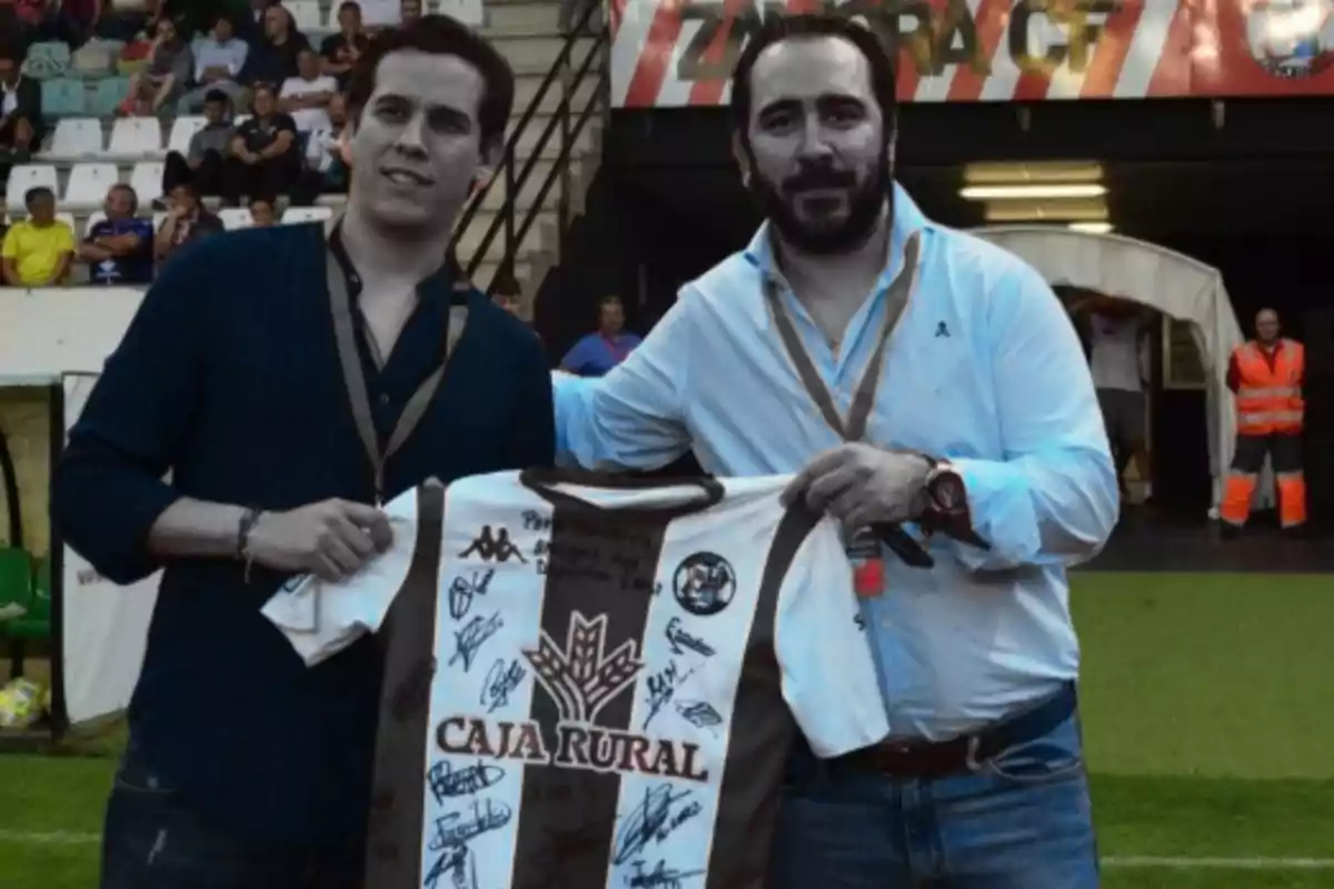 Dos hombres posan en un estadio de fútbol sosteniendo una camiseta firmada del equipo Zamora CF mientras sonríen a la cámara