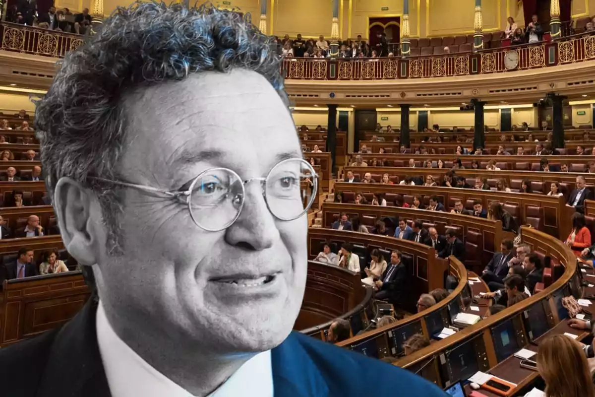 A man with glasses in the foreground against a background of a parliament full of people.