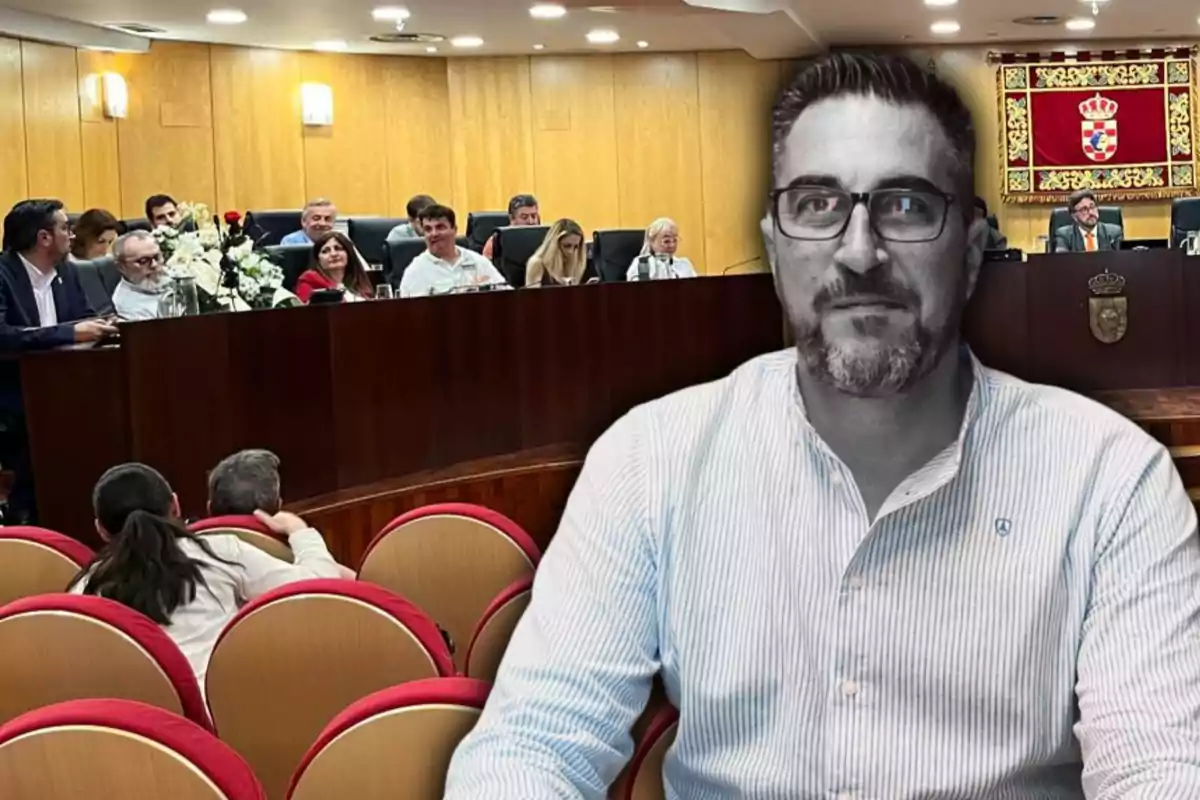 Un hombre con gafas en primer plano aparece en blanco y negro frente a una sala de reuniones con varias personas sentadas en una mesa larga y público en butacas rojas, al fondo hay un escudo oficial en la pared.
