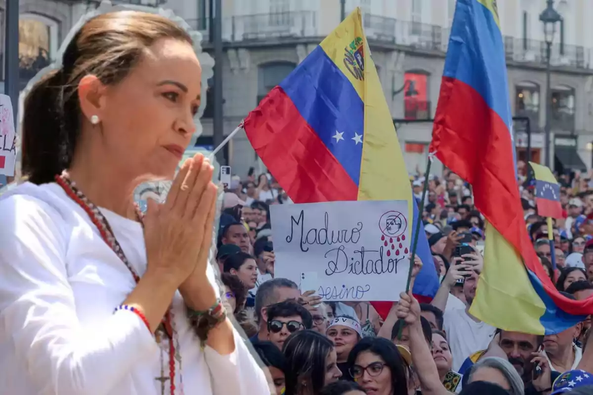 Una mujer con las manos juntas en actitud de oración aparece en primer plano, mientras al fondo se observa una multitud de personas con banderas de Venezuela y carteles de protesta.