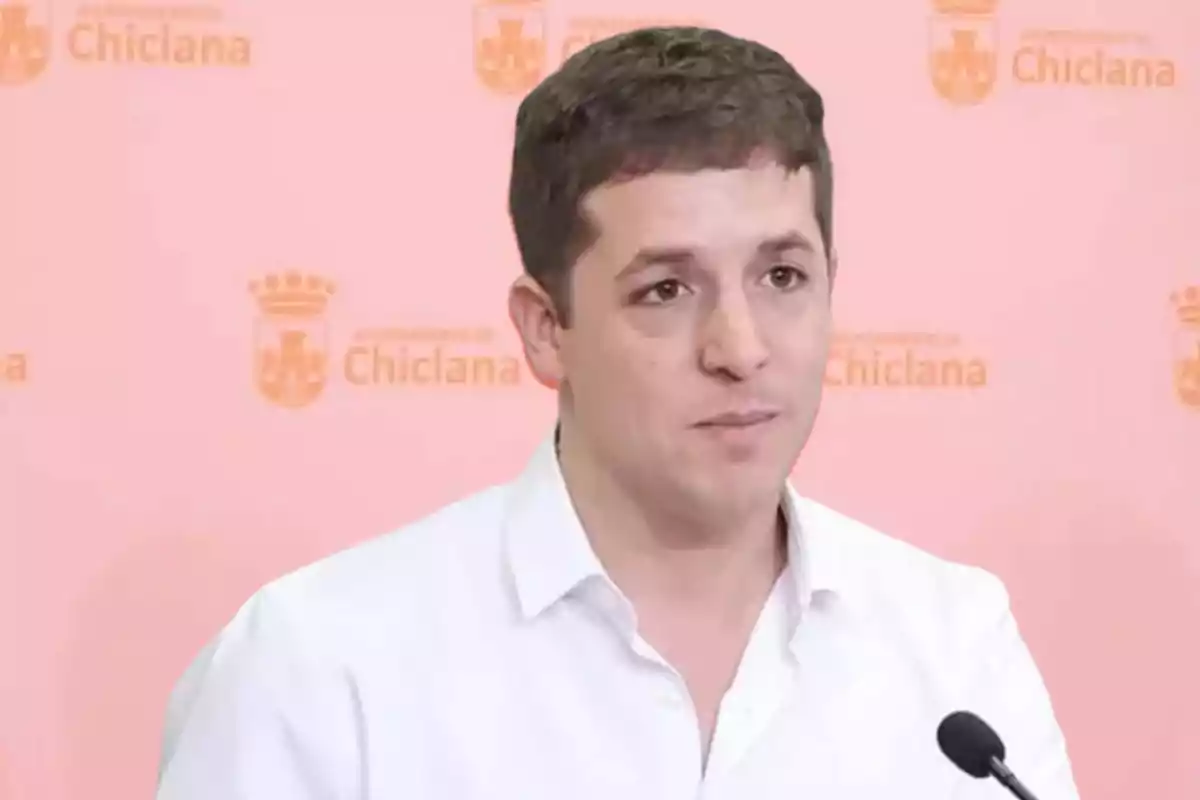 Un hombre con camisa blanca hablando frente a un micrófono con un fondo que tiene el logotipo del Ayuntamiento de Chiclana.