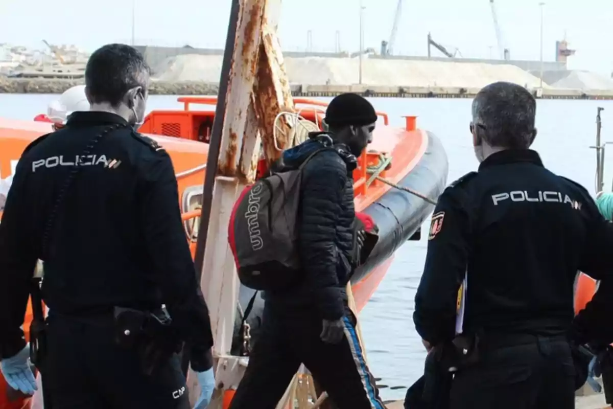 Un grupo de policías junto a un hombre con mochila cerca de una embarcación en un puerto.