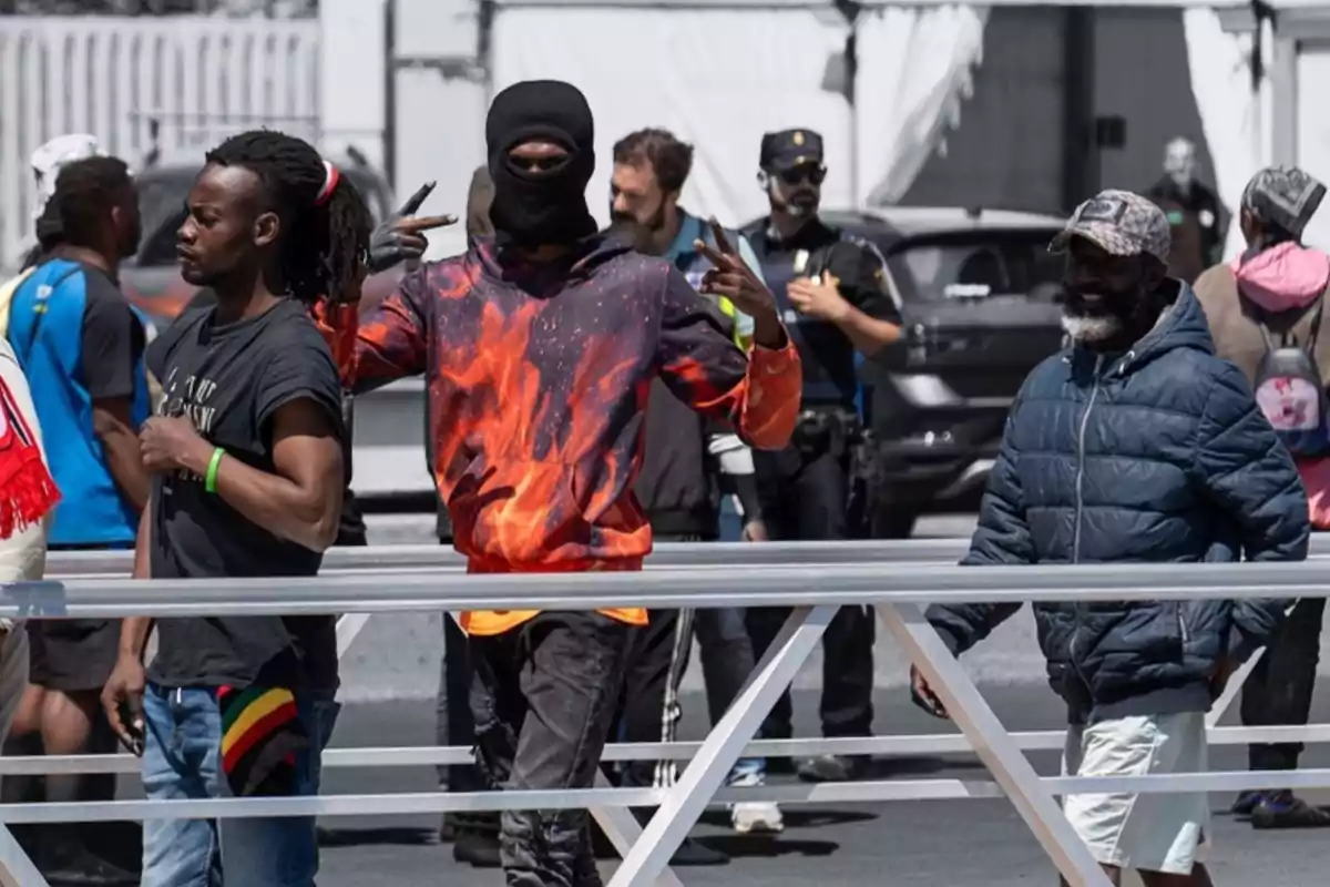 Un grupo de personas camina por la calle, una de ellas lleva una sudadera con diseño de llamas y hace un gesto con las manos, mientras que al fondo se observa a un policía.