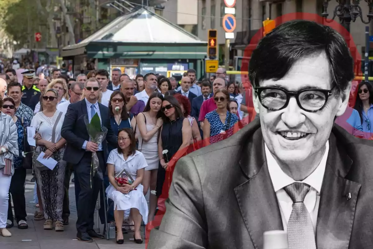 Una multitud de personas se reúne en una calle, algunas con flores blancas, mientras en primer plano se ve una imagen en blanco y negro de un hombre con gafas y traje.