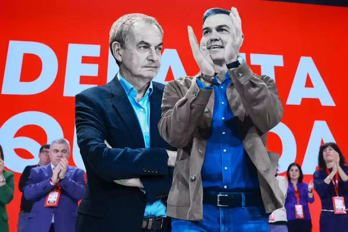 Dos hombres de pie en un escenario con fondo rojo, uno con los brazos cruzados y el otro aplaudiendo, rodeados de personas que también aplauden.