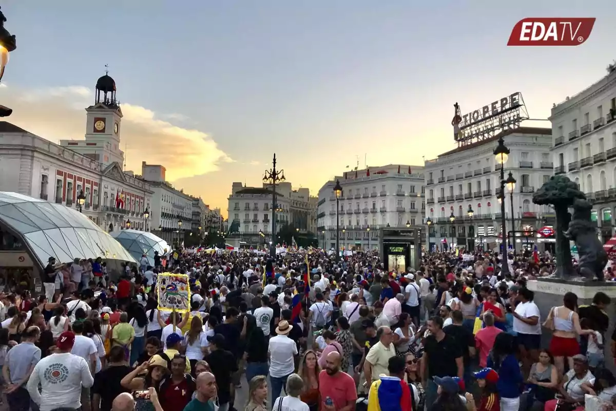 Una multitud de personas se reúne en una plaza histórica al atardecer, con edificios emblemáticos y un reloj visible en el fondo.