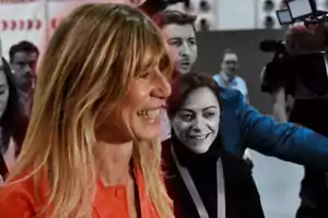 Una mujer sonriente con cabello rubio en primer plano mientras otras personas están en el fondo, algunas en blanco y negro.