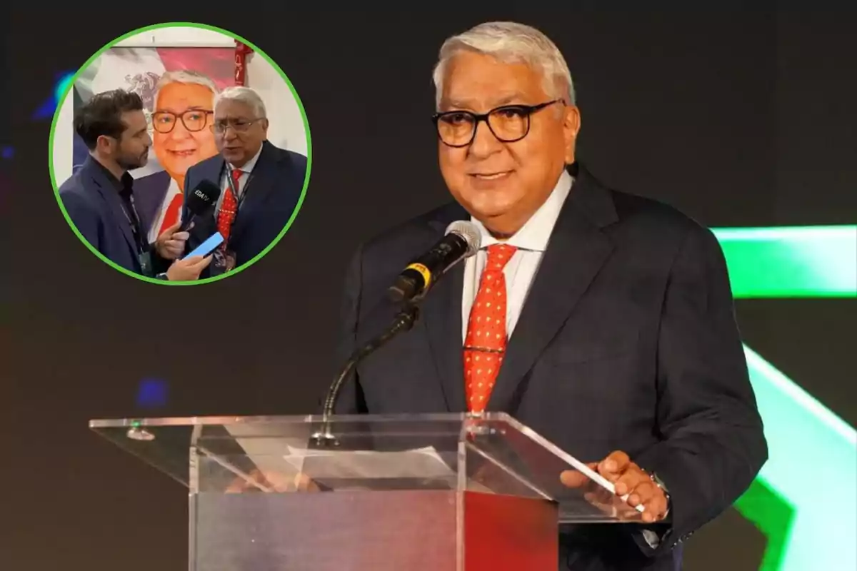 Un hombre de cabello canoso y gafas, vestido con un traje oscuro y corbata roja, está hablando en un podio con un micrófono; en la esquina superior izquierda hay un círculo verde que muestra una imagen de él siendo entrevistado por otra persona.