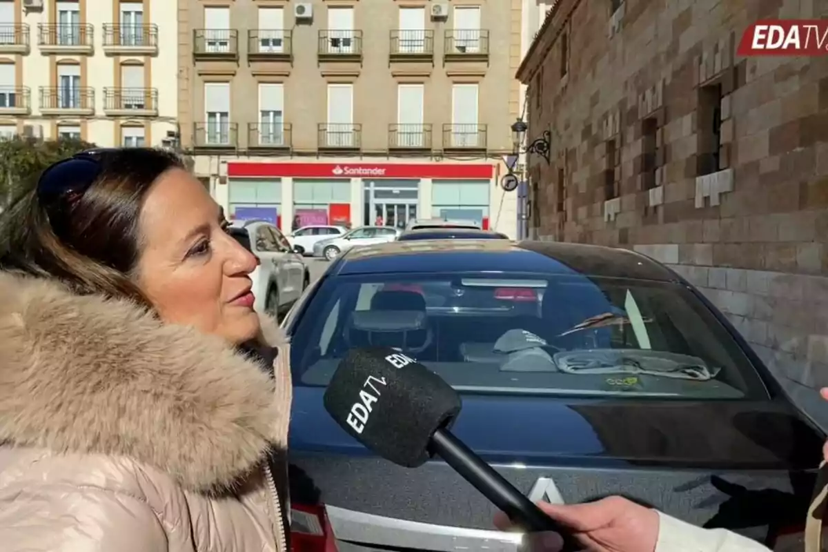 A woman in a fur coat is being interviewed on the street by a reporter holding an EDATV microphone, with a car and a building in the background.