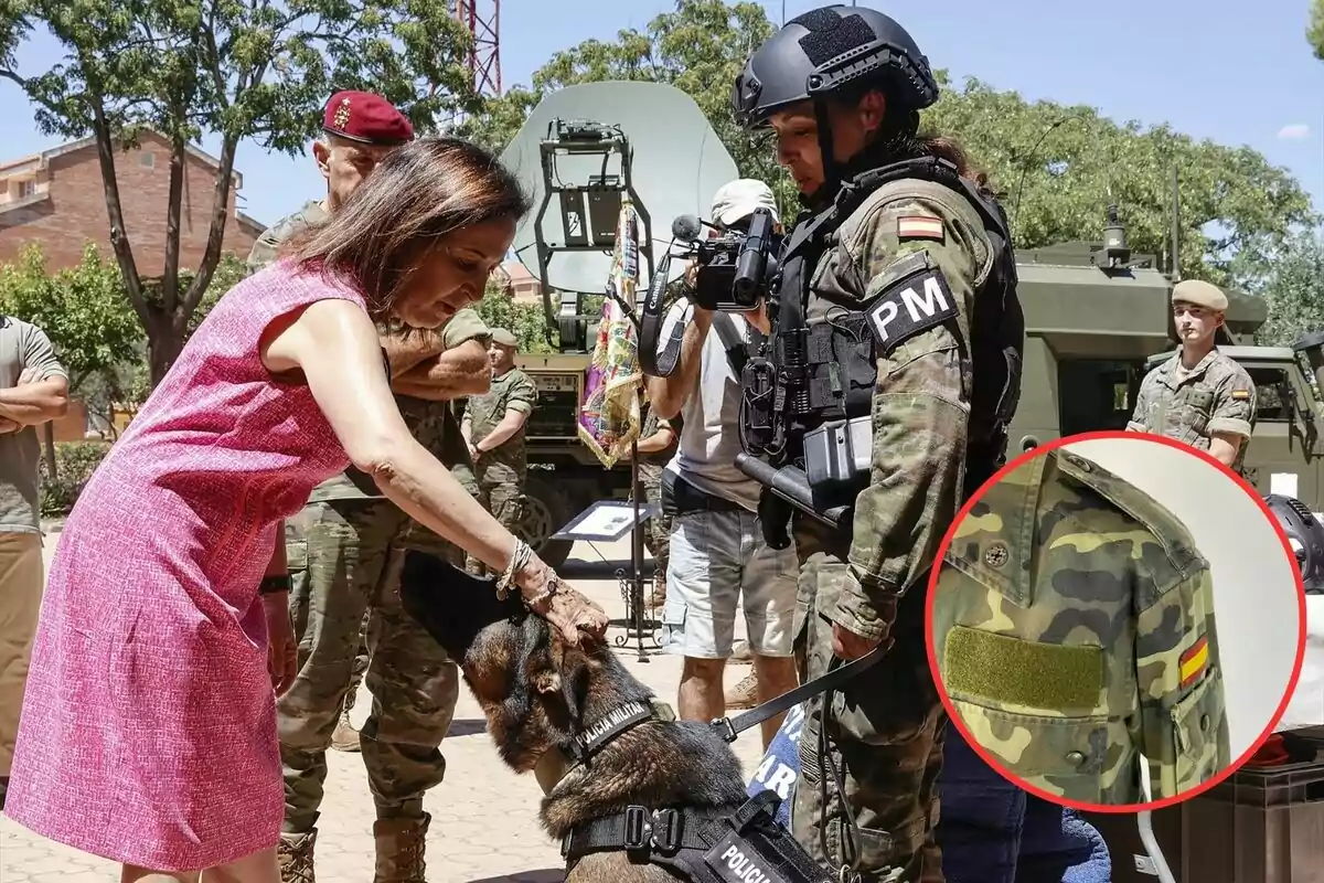 Una mujer vestida de rosa acaricia a un perro policía mientras un soldado con uniforme de camuflaje y casco observa, con otros militares y equipo militar en el fondo.