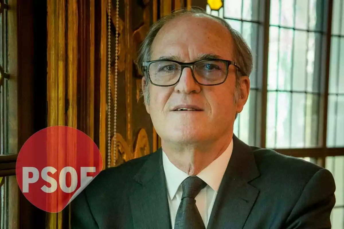 Un hombre con gafas y traje oscuro posa frente a una ventana con el logo del PSOE en la esquina inferior izquierda.