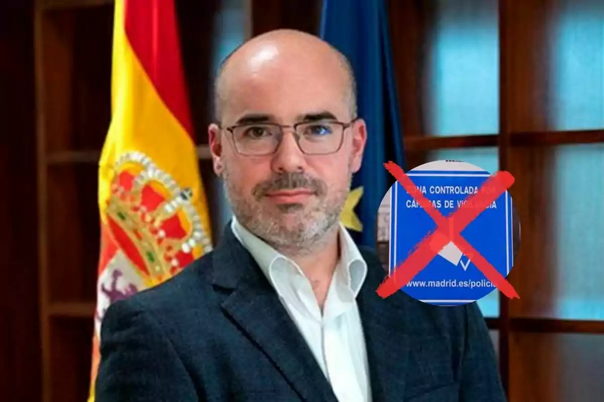 A man with glasses and a beard, dressed in a suit, is standing in front of a Spanish flag and another blue flag, with a blue sign crossed out with a red X next to him.