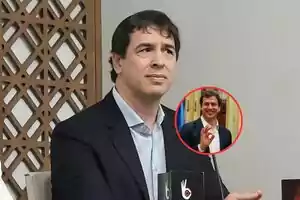 Un hombre con traje oscuro y camisa clara está sentado frente a un micrófono con un diseño decorativo en la pared detrás de él y un círculo rojo muestra una imagen más pequeña de él sonriendo y haciendo un gesto con la mano.