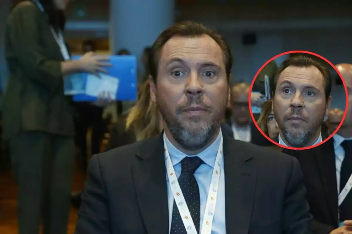 Un hombre con traje y corbata está sentado en un evento, con una expresión seria; en el fondo, otras personas están presentes y una mujer sostiene documentos.