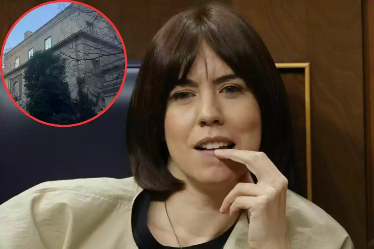 Una mujer con cabello corto y oscuro se lleva la mano a la boca mientras está sentada, con una imagen de un edificio en un círculo rojo en la esquina superior izquierda.