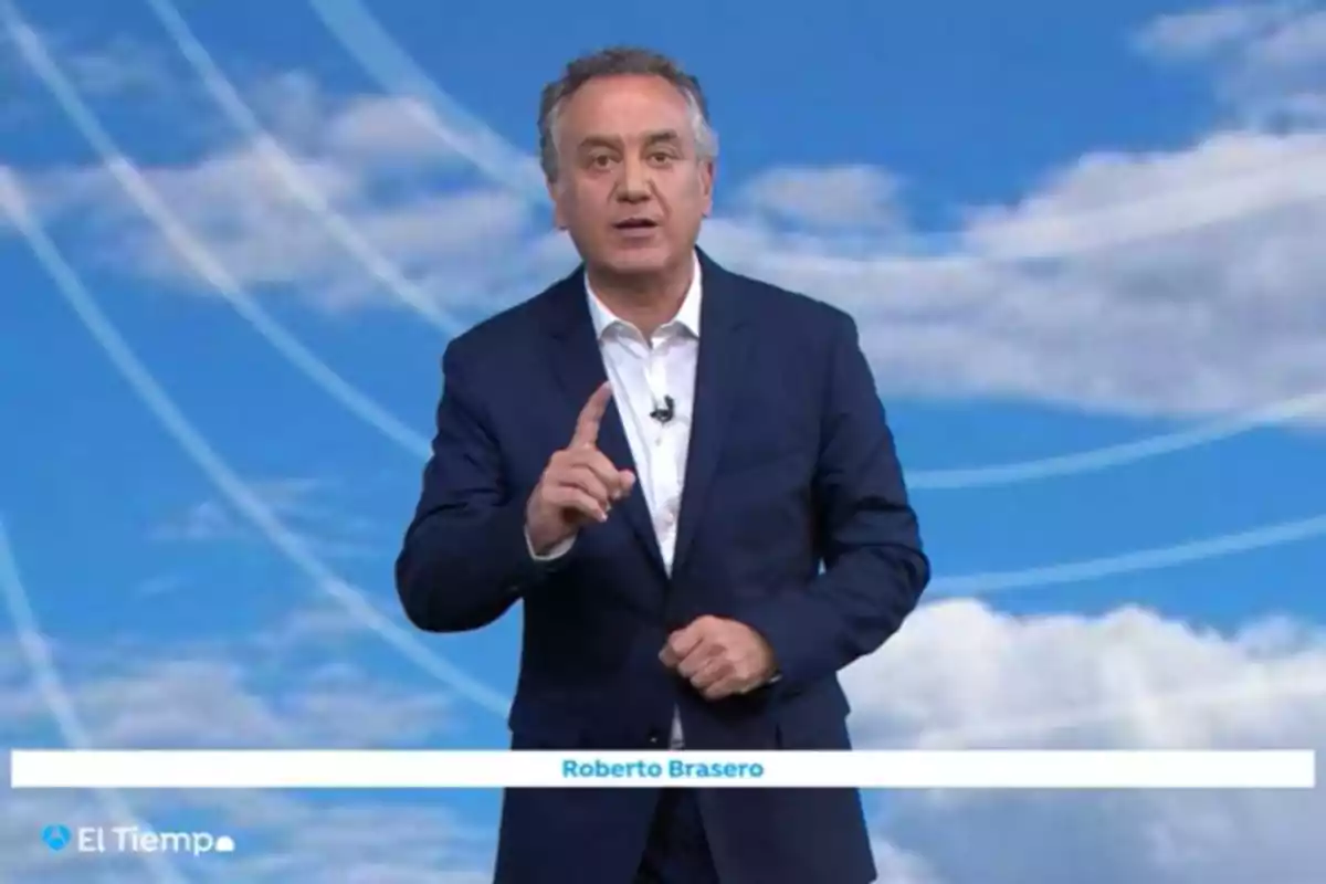 Un hombre en traje oscuro está de pie frente a un fondo azul con nubes y líneas blancas, gesticulando con una mano mientras habla. Un hombre en traje oscuro está de pie frente a un fondo azul con nubes y líneas blancas, gesticulando con una mano mientras habla.