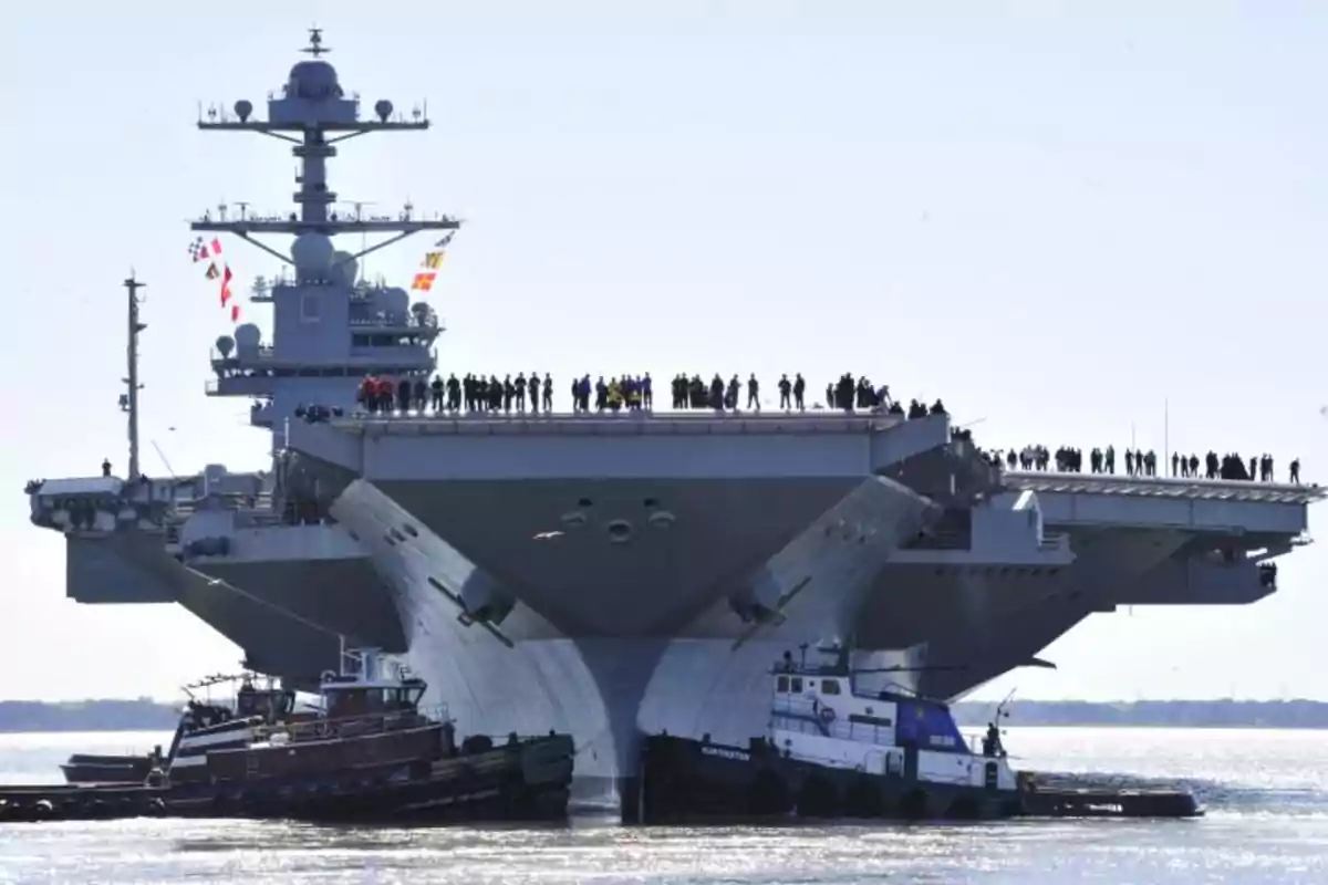 Un portaaviones con varias personas en la cubierta y remolcadores a su alrededor.