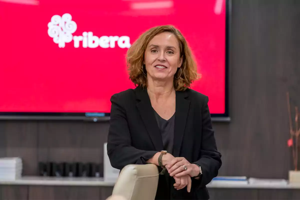 A woman with brown hair and a dark suit is standing in front of a red screen with the "ribera" logo.