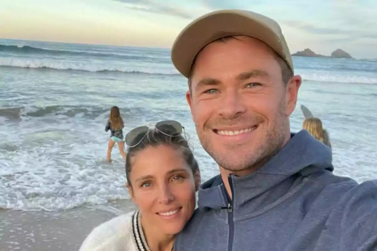 Una pareja, formada por Elsa Pataky y Chris Hemsworth, sonriente tomándose una selfie en la playa con dos niñas jugando cerca del mar al atardecer. Una pareja, formada por Elsa Pataky y Chris Hemsworth, sonriente tomándose una selfie en la playa con dos niñas jugando cerca del mar al atardecer.