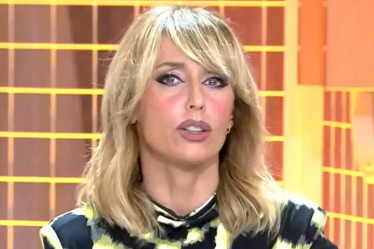 Emma García con cabello rubio y maquillaje pronunciado en un Fiesta con fondo de líneas naranjas.