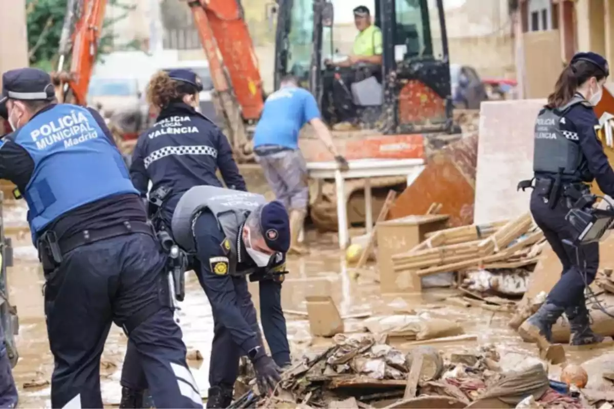 Policías municipales y locales colaboran en la limpieza de escombros tras una inundación, con maquinaria pesada en el fondo.