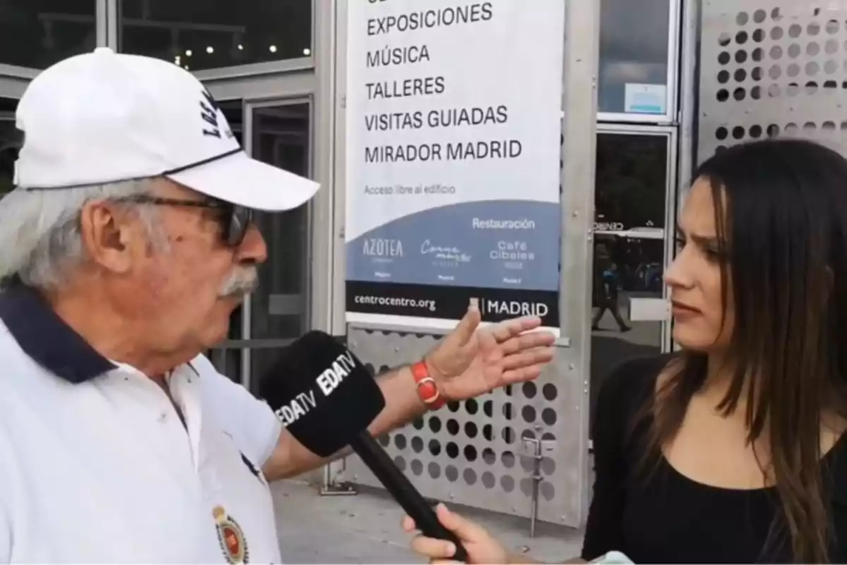 Un hombre mayor con gorra blanca y gafas de sol es entrevistado por una reportera frente a un edificio con un cartel que anuncia exposiciones, música, talleres, visitas guiadas y un mirador en Madrid.