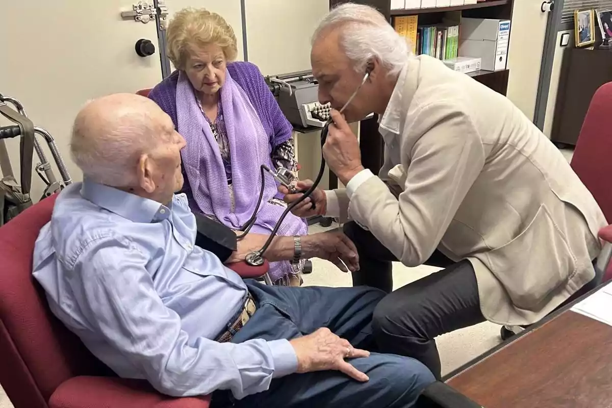 Un médico revisa a un paciente mayor con un estetoscopio mientras una mujer mayor observa.