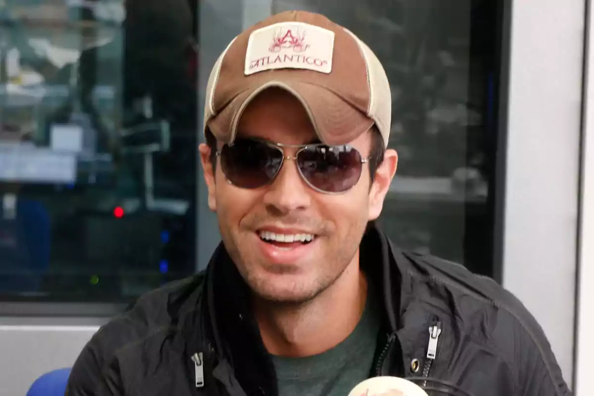 Enrique Iglesias con gorra y gafas de sol sonríe mientras sostiene un micrófono. Enrique Iglesias con gorra y gafas de sol sonríe mientras sostiene un micrófono.